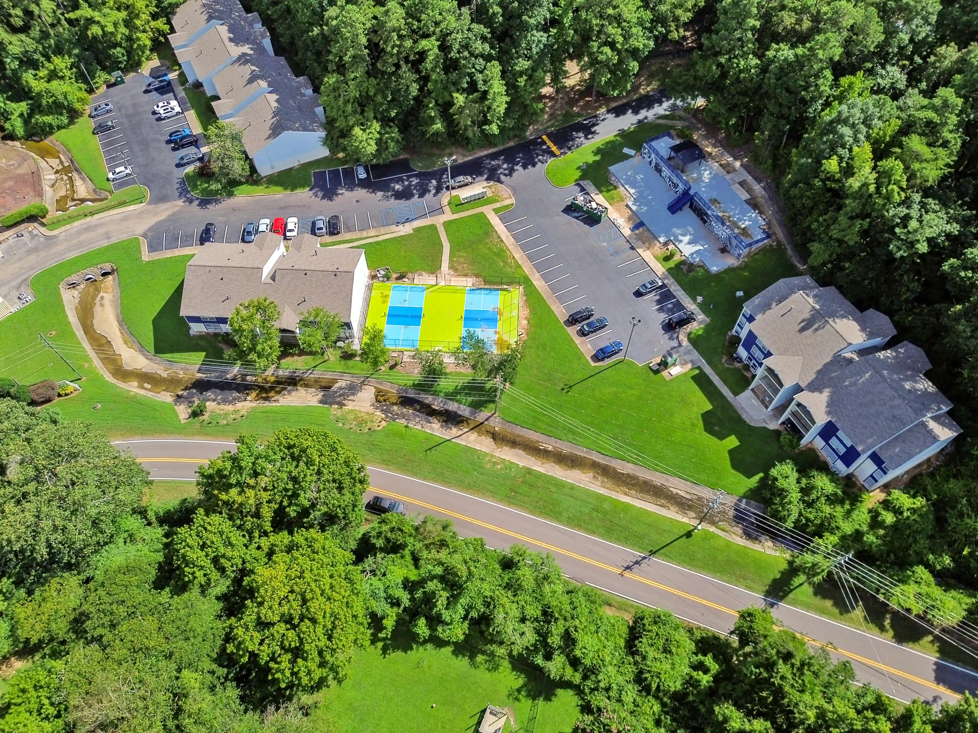 Aerial drone view of an apartment community featuring residential buildings, parking areas, green spaces, and pickleball courts.