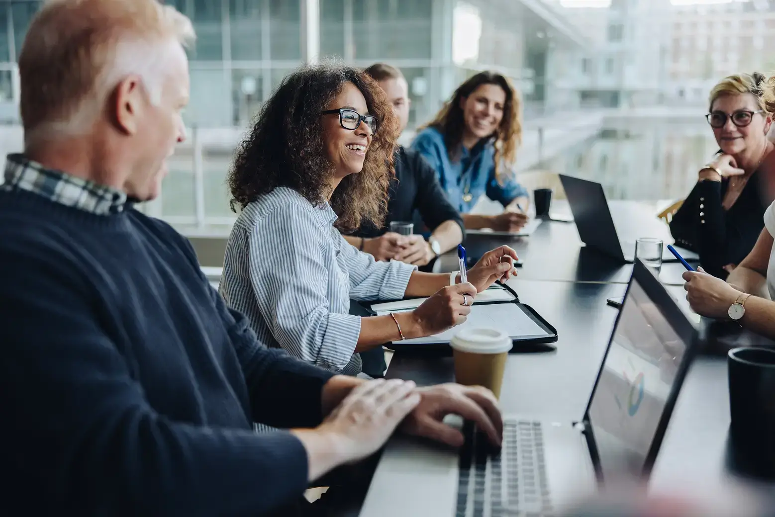 Diverse team of professionals collaborating in a modern office meeting with laptops, notebooks, and coffee in a bright conference space.