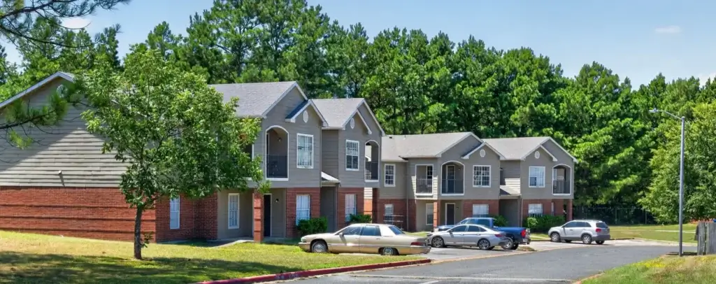 Multi-unit apartment buildings with brick and siding exteriors, landscaped lawns, parking spaces, and mature pine trees.