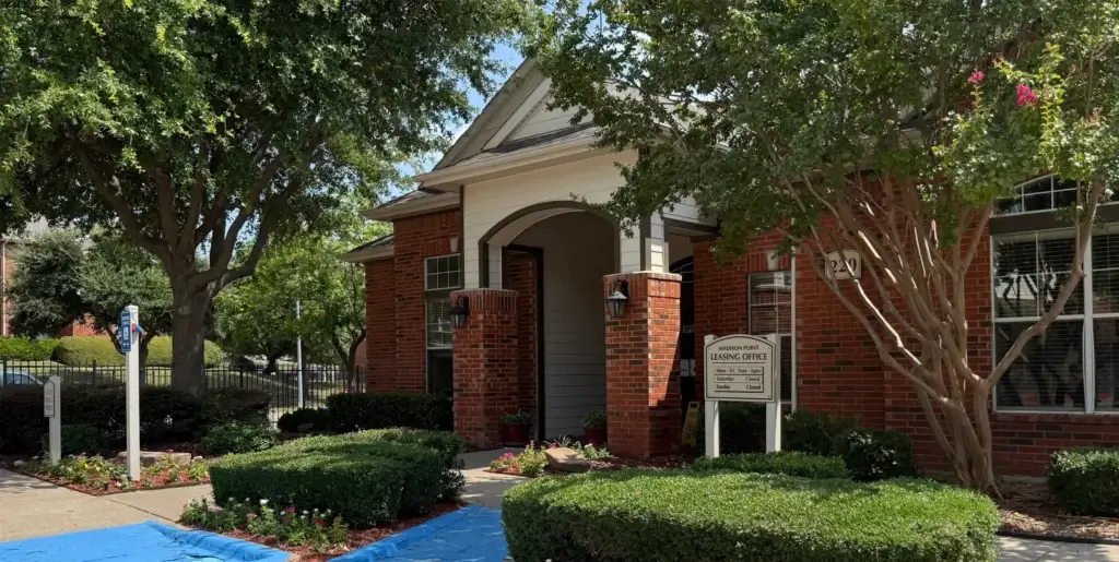 Brick apartment community leasing office with landscaped grounds, accessible parking, and a blue-painted walkway.