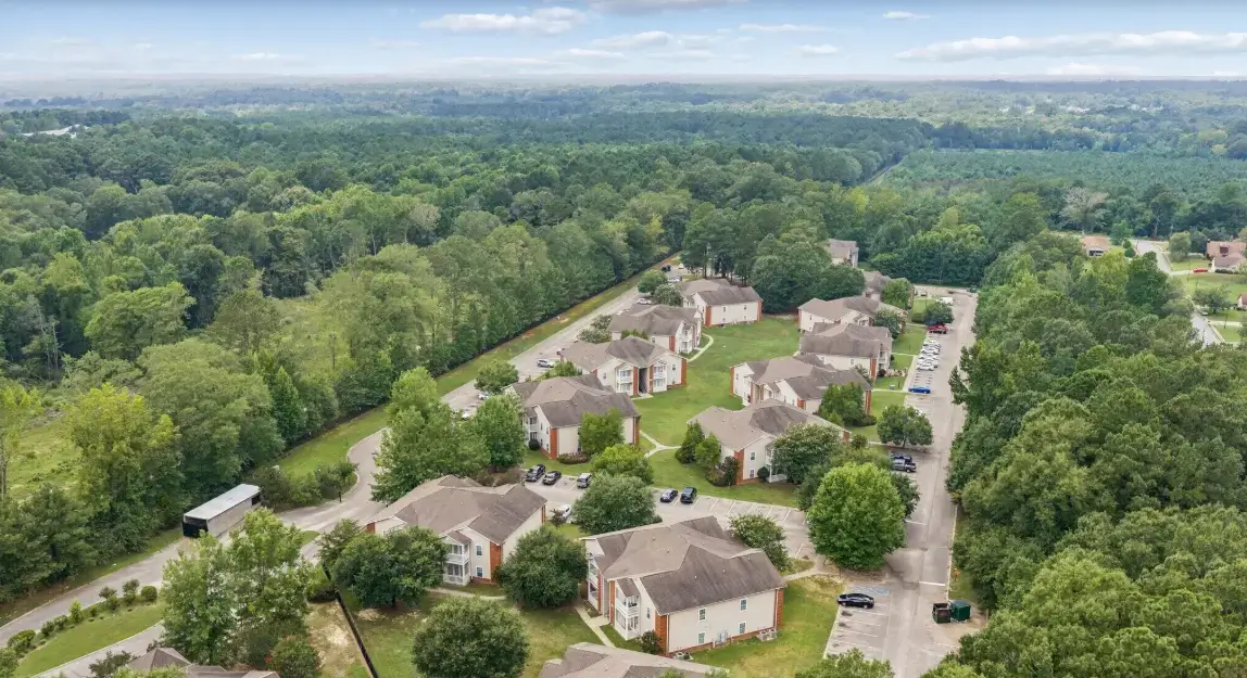 Aerial view of an apartment community surrounded by dense trees, featuring multiple residential buildings, landscaped grounds, roads, and parking areas.