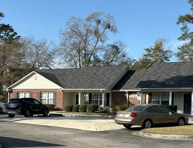 Single-story brick apartment building with a covered entrance, landscaped shrubs, parking area, and mature trees.