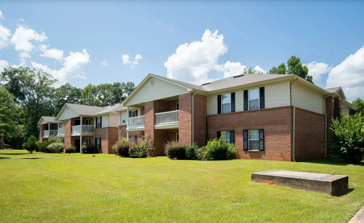 Brick apartment buildings with balconies, landscaped lawns, shrubs, and mature trees under a bright blue sky.