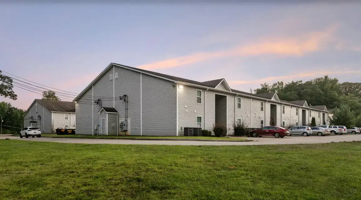 Exterior view of a gray apartment building with a large grassy lawn, parking area, parked cars, and trees at dusk.