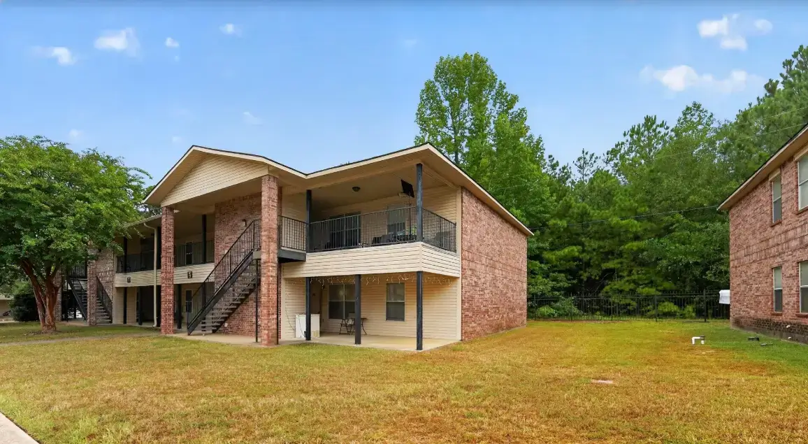 Exterior view of an apartment building at Emerald Trace and Cinnamon Court featuring brick walls, covered balconies, outdoor stairs, grassy grounds, and mature trees.