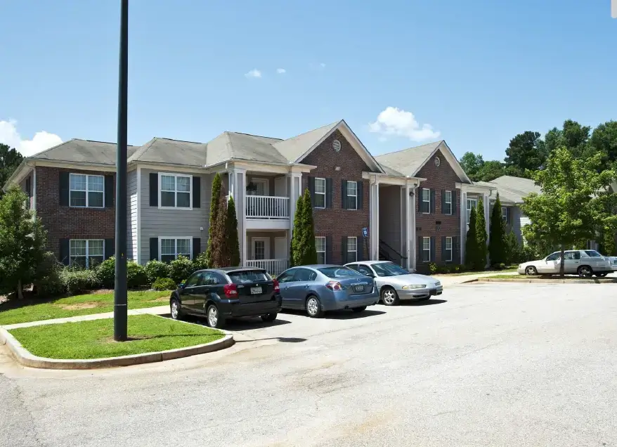 Brick apartment community with balconies, landscaped grounds, parking spaces, and mature trees under a clear blue sky.