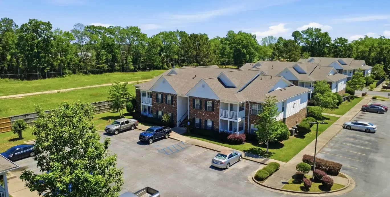 Aerial view of a multi-building apartment community with brick exteriors, balconies, landscaped grounds, parking lots, and surrounding trees.
