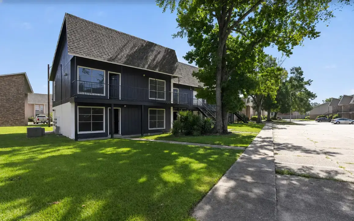 Exterior view of Magnolia Trace apartments featuring a dark two-story building, balconies, landscaped lawn, mature trees, sidewalks, and parking.