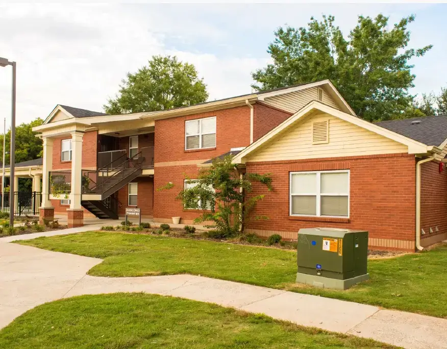Exterior view of a Spring Branch apartment building featuring red brick construction, covered balconies, landscaped lawns, concrete walkways, and mature trees.