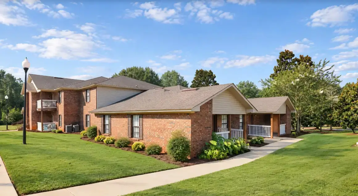 Exterior view of Susanna Ridge apartments featuring red brick buildings, covered porches, landscaped lawns, concrete walkways, and mature trees.