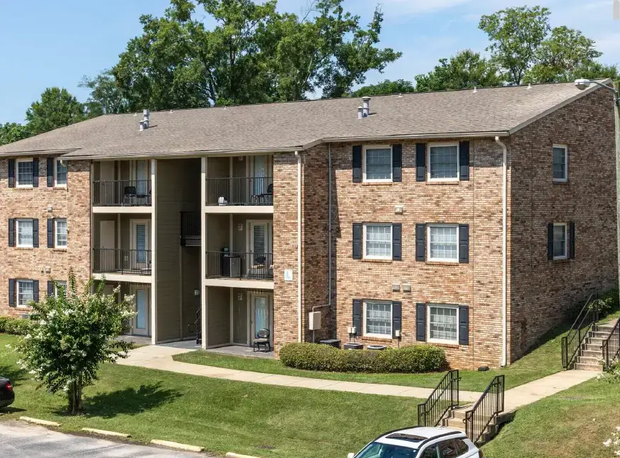 Exterior view of The Hill Apartments featuring a three-story brick building, covered balconies, landscaped lawn, walkways, and outdoor stairs.