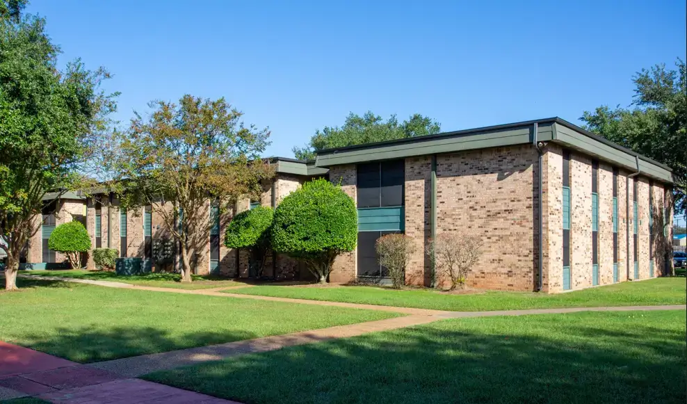 Exterior view of The Melrose apartment building with brick walls, landscaped lawns, trimmed shrubs, mature trees, and concrete walkways.